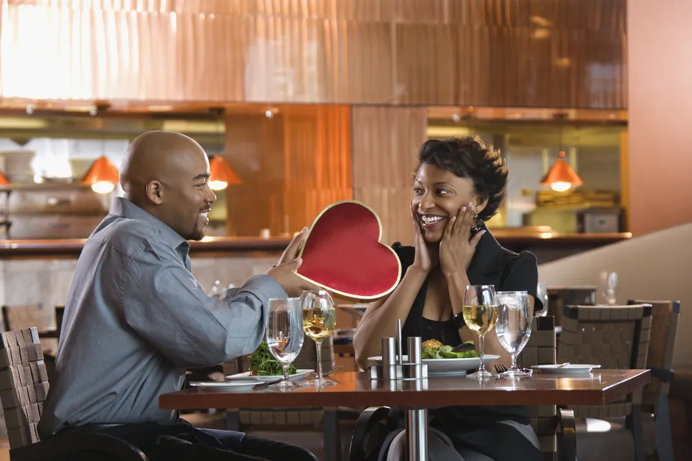 Couple at a restaurant table, the man surprising the woman with a red heart-shaped box
