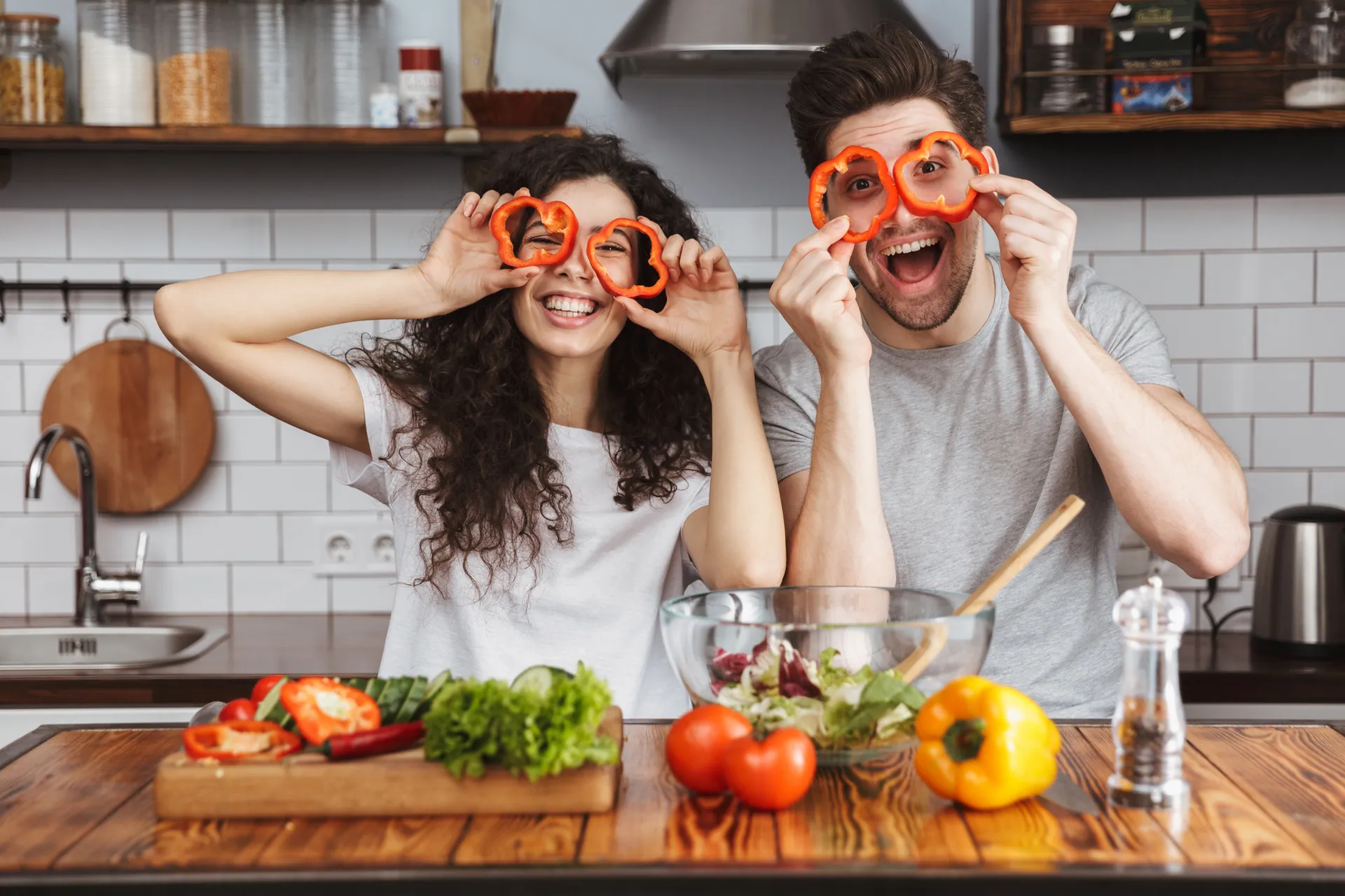 Couple in a kitchen holding pepper rings up to their eyes like glasses