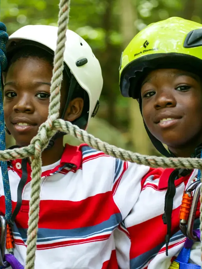 Two smiling boys in climbing helmets look through a rope net on the ropes course