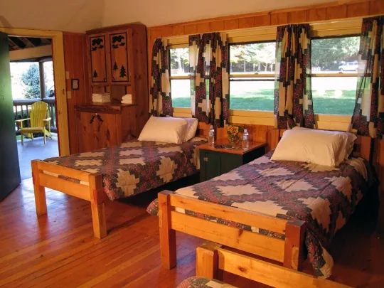 Cabin room with two wooden beds under patterned quilts and windows looking out on the lawn