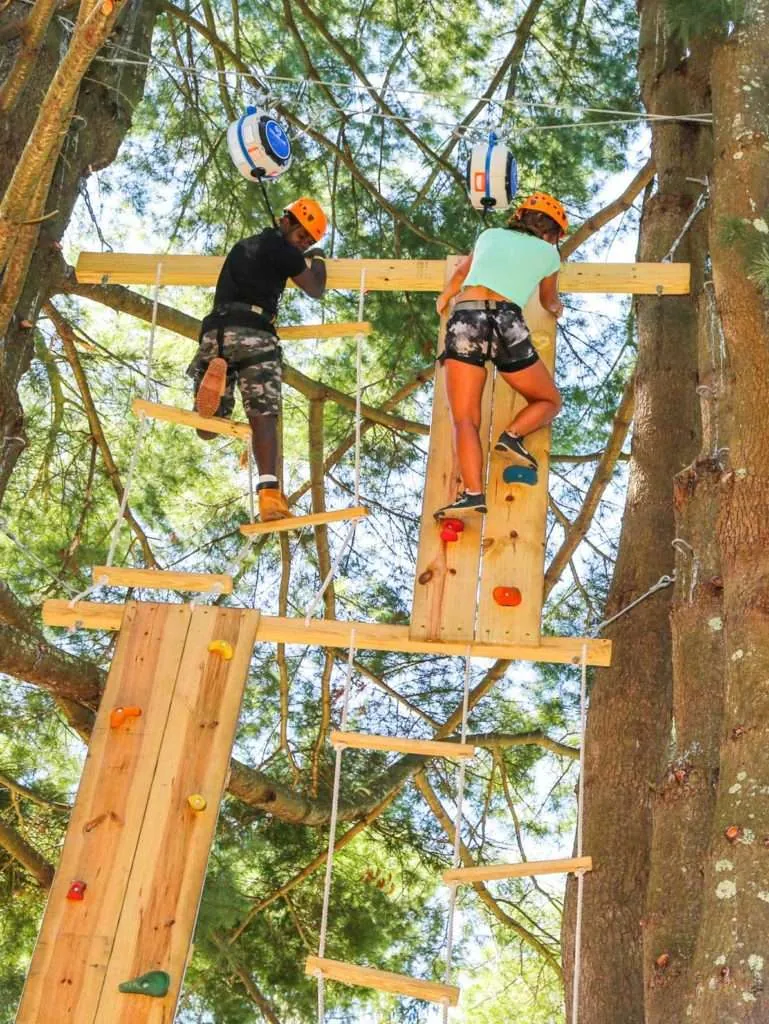 Two kids in helmets climb side by side up the wooden high ropes course among tall trees