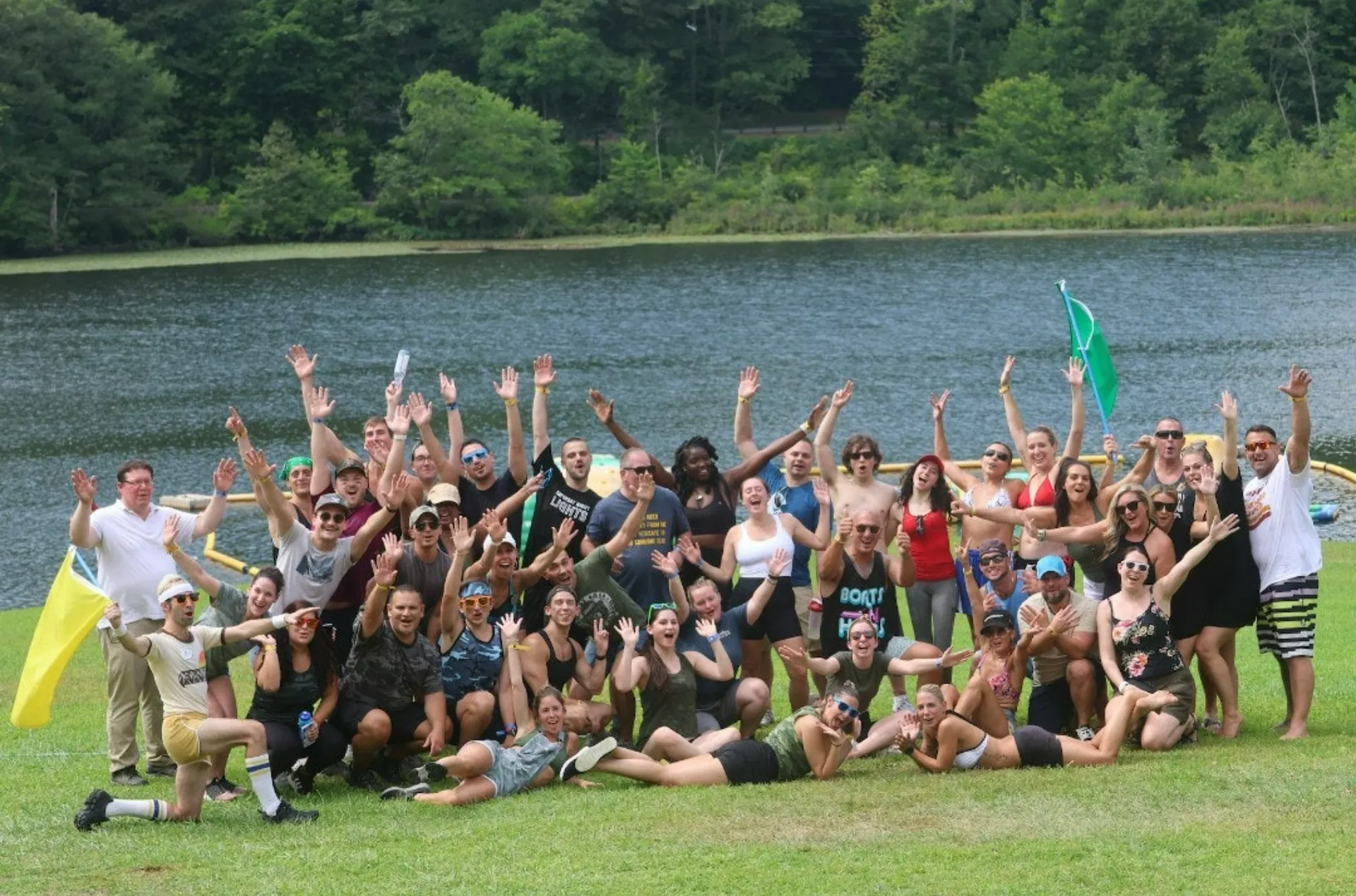A large group of guests cheering with arms raised on the lawn in front of the lake
