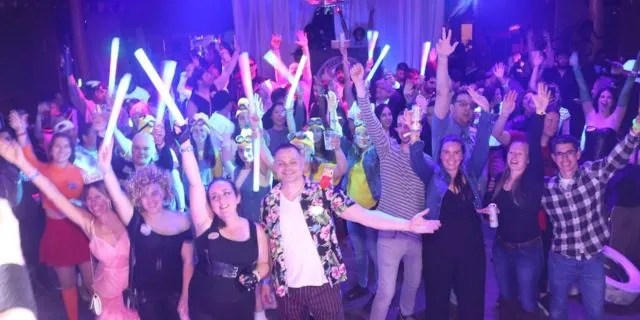 A crowd of costumed guests waving glow sticks and cheering at a purple-lit party