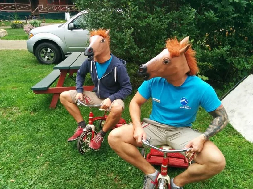 Two crew members in horse-head masks riding tiny tricycles on the lawn