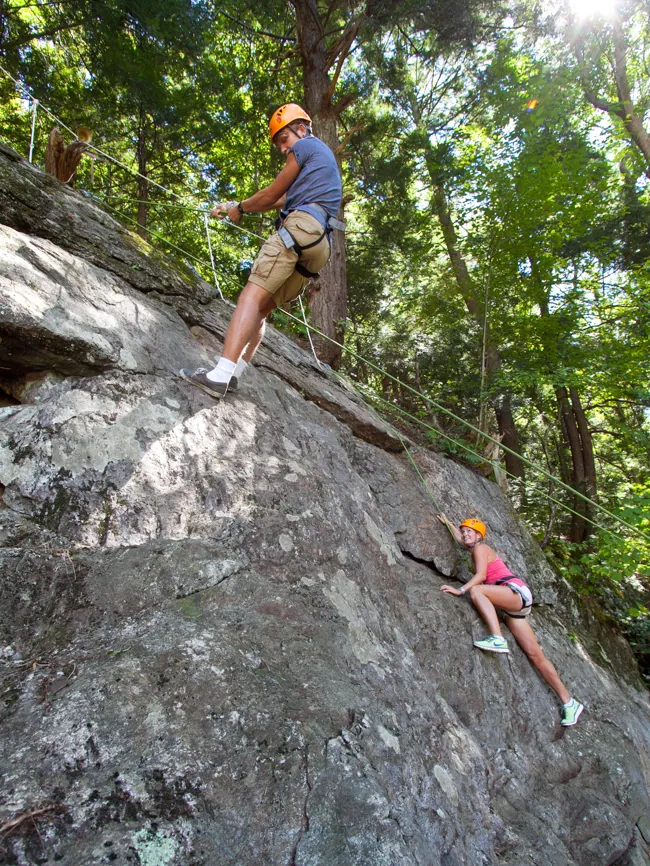 Two climbers in helmets and harnesses scaling a large rock face in the woods