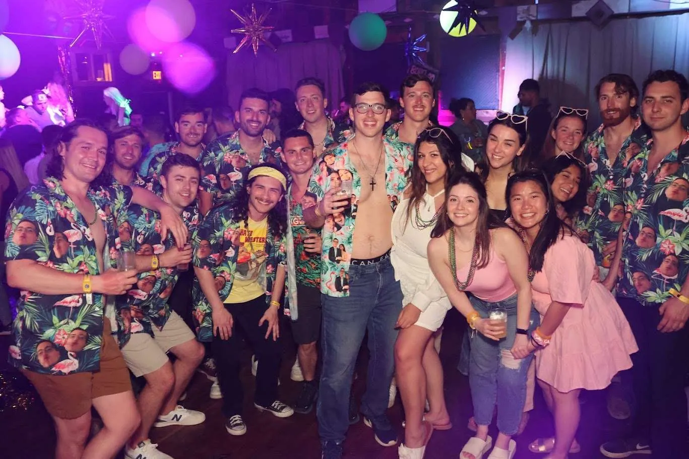 A crowd of guests in Hawaiian shirts and party outfits posing together under purple lights at a themed party