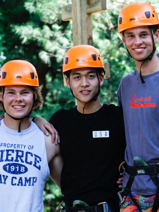 Three young men in orange climbing helmets and harnesses smiling on the ropes course