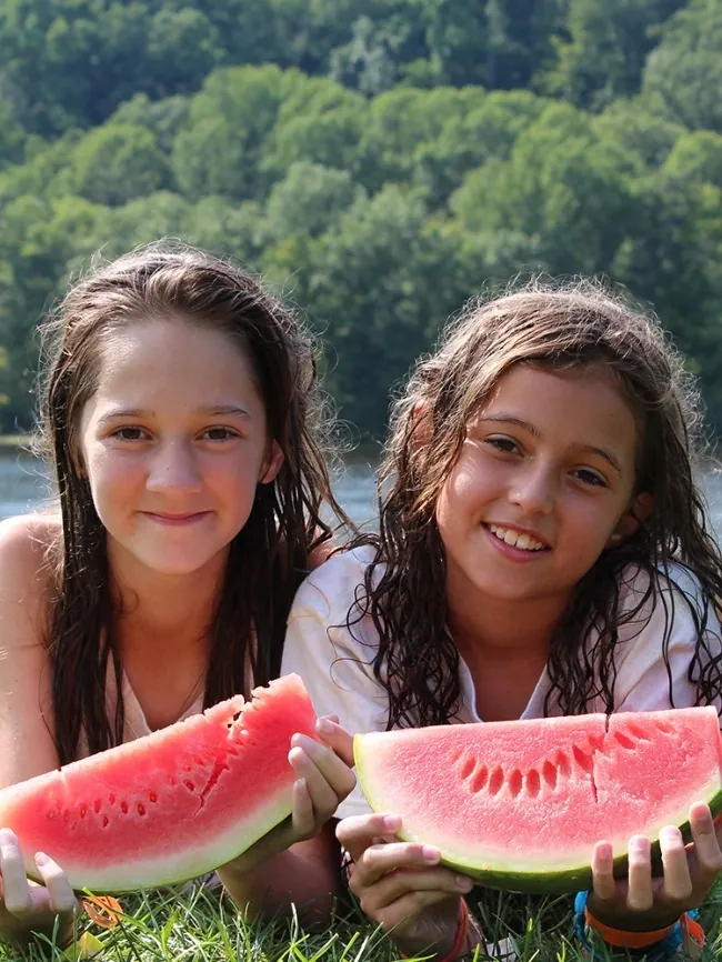 Two smiling girls lying on the grass holding big slices of watermelon by the lake