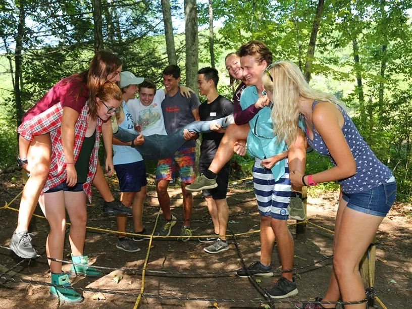 Teens laughing as they carry a teammate through a rope challenge in the woods
