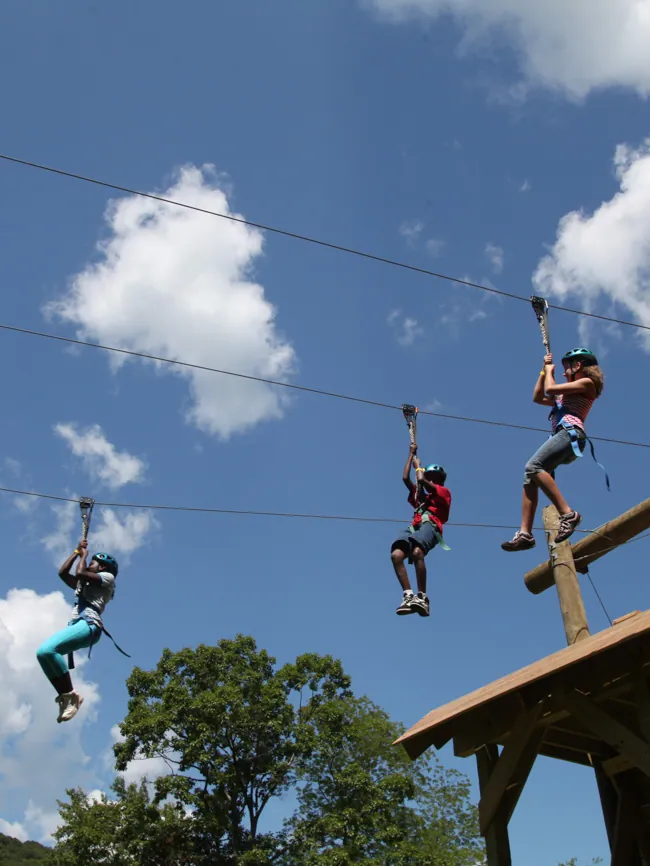 Three guests race side by side on parallel zip lines under a blue sky