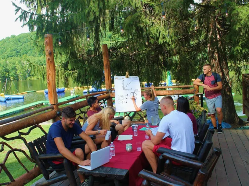 Team meeting around a table on a wooden deck by the lake, with one person presenting at a flip chart