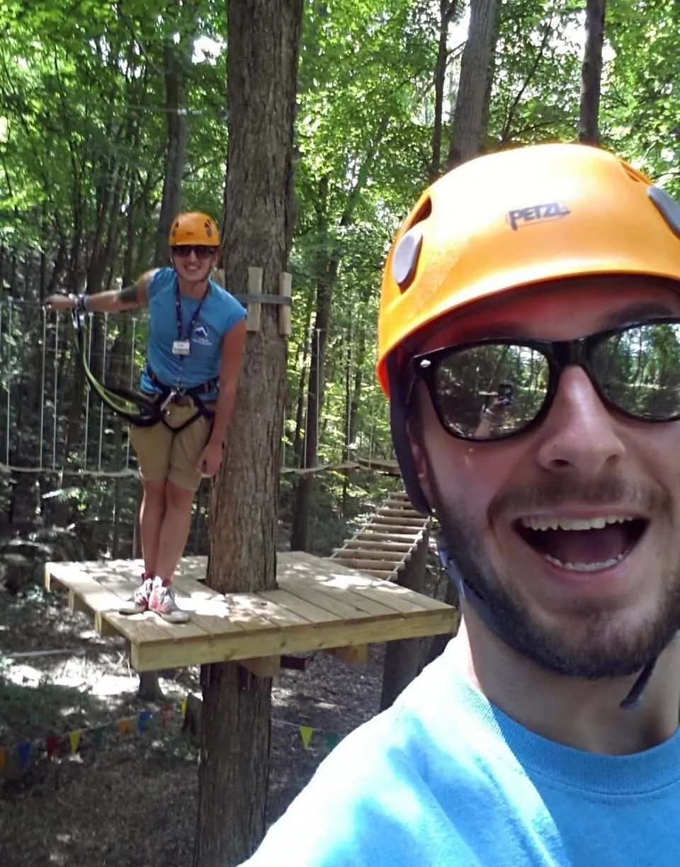 Crew member in an orange helmet taking a selfie below a teammate standing on a treetop platform