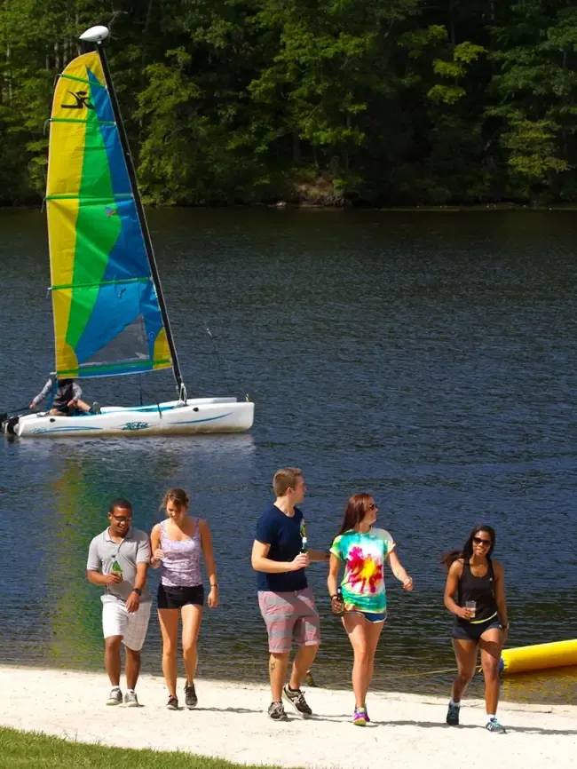 Five people walking on a sandy lakeshore with a colorful sailboat on the calm water behind them