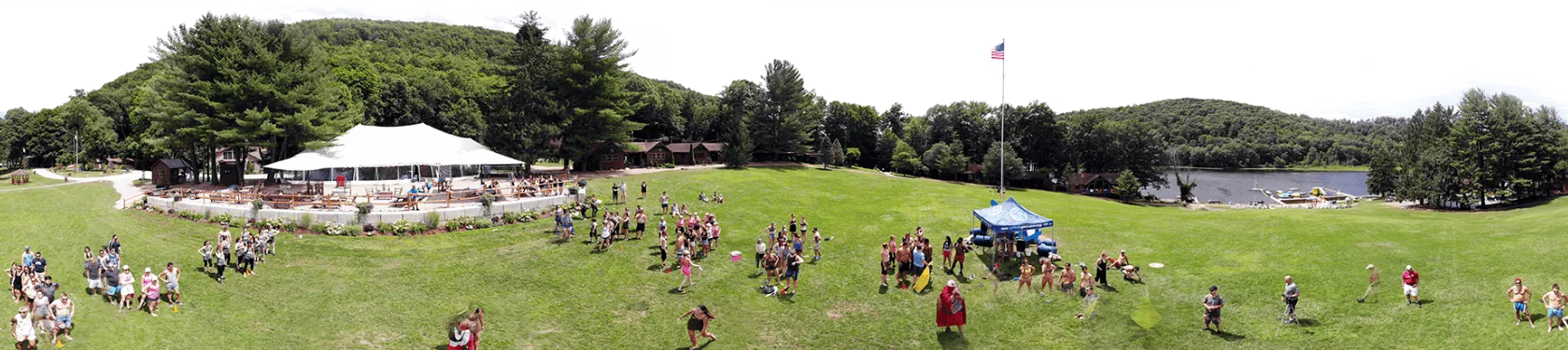 Panoramic view of camp: the lawn, a white tent, the flagpole and the lake ringed by trees