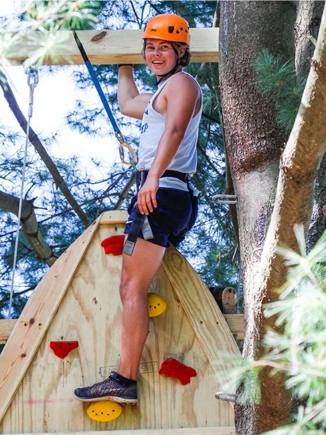 A smiling teen in an orange helmet and harness climbs a wooden wall on the ropes course