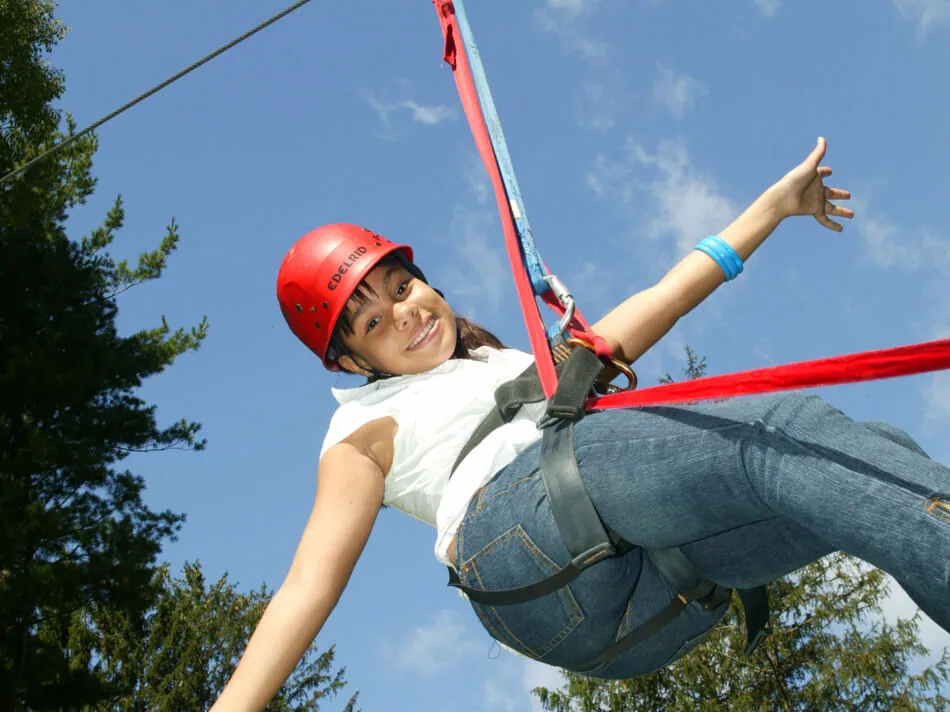 A smiling guest in a red helmet leans back in her harness, arms out, on a zip line against a blue sky