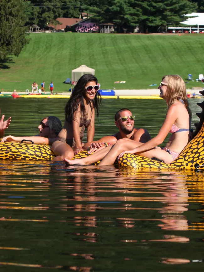 Friends lounging on a yellow dragon-shaped float on the lake, with the camp lawn behind