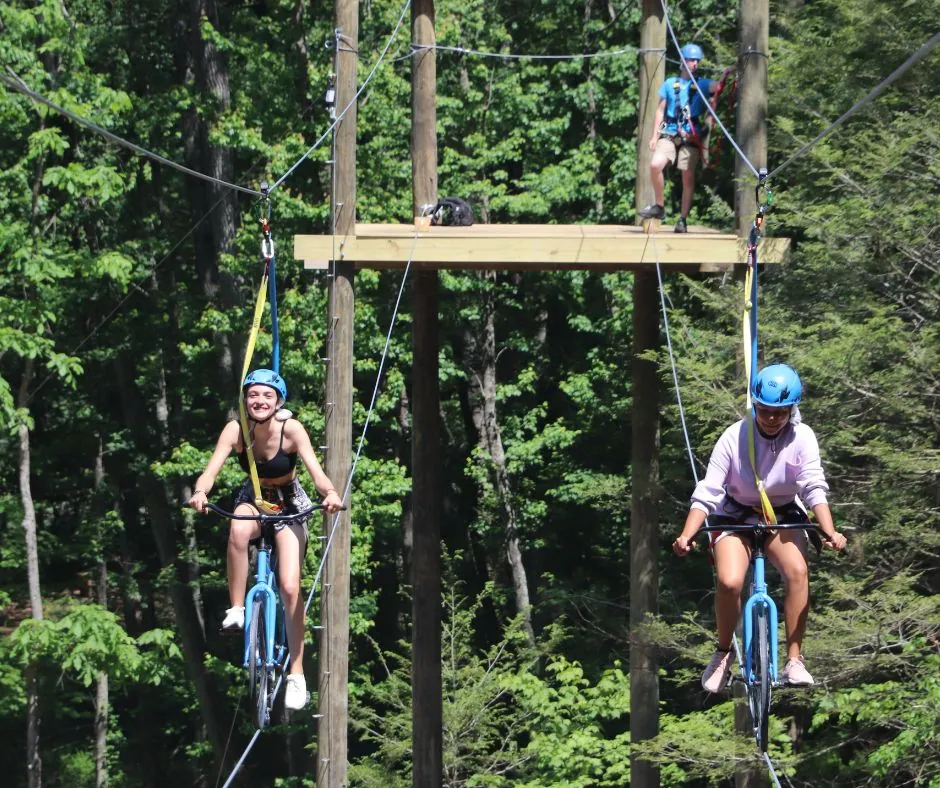 Two guests in helmets pedal bicycles along a high wire between trees on the aerial course