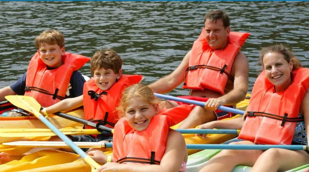 A smiling family in orange life jackets paddles yellow kayaks on the lake