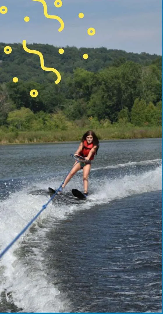 A girl in a red life vest waterskis across the lake with wooded hills behind her